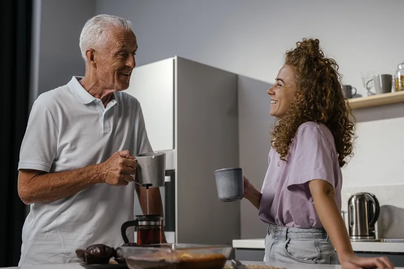 Un hombre mayor y una mujer joven toman un café y se ríen en una cocina.