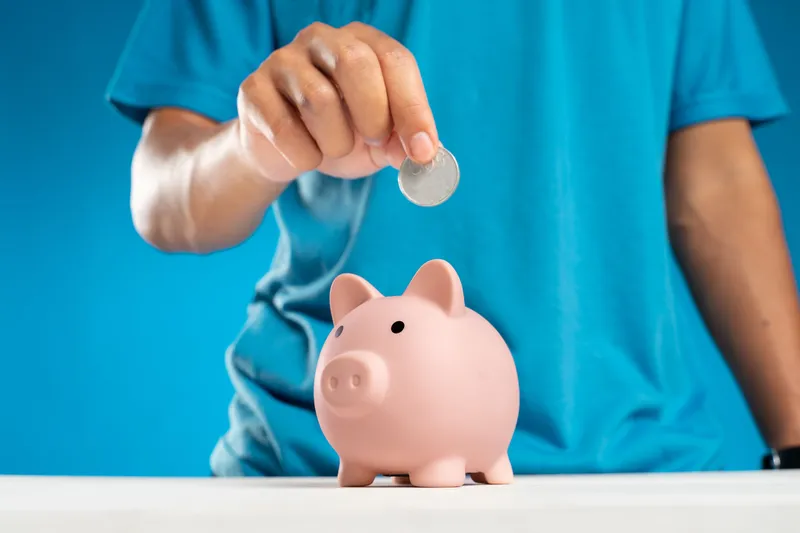 Una mano echa una moneda en una hucha de cerdito rosa.